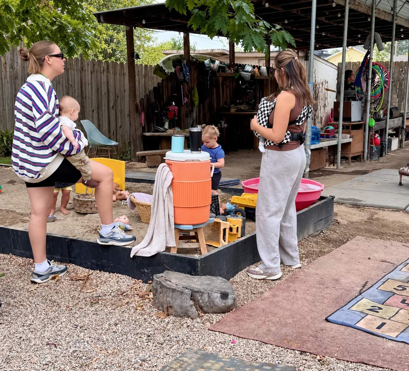Two caregivers standing beside the sand play area with their children