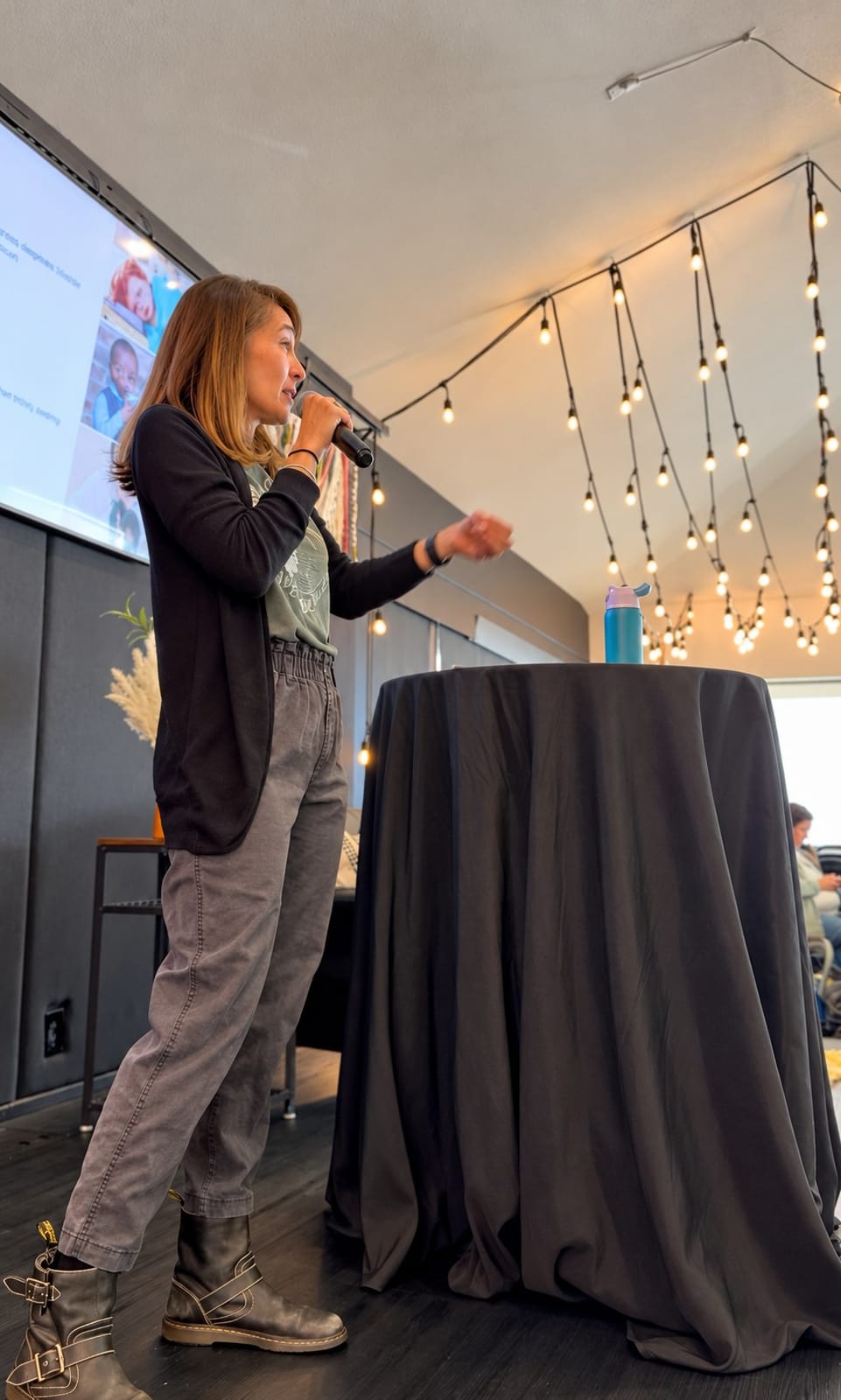 Daniele standing onstage in jeans, speaking into a microphone beneath string lights